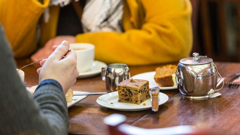 A cake and metal tea pot on top of a wooden table. Two people sit around the table.